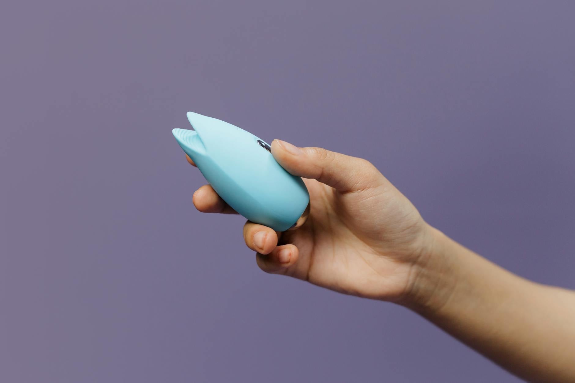Two hands holding pink and blue silicone vibrators together against a pastel background